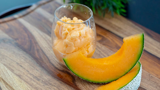 Two cantaloupe are placed next to a clear cup filled with light orange cantaloupe slushy.