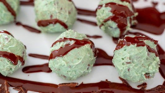 Six green doughy balls are aligned in two rows of three. Chocolate sauces is drizzled across the green dough balls. Some of the chocolate is on the white surface below the balls. These are a bite of healthy's mint chocolate truffles.