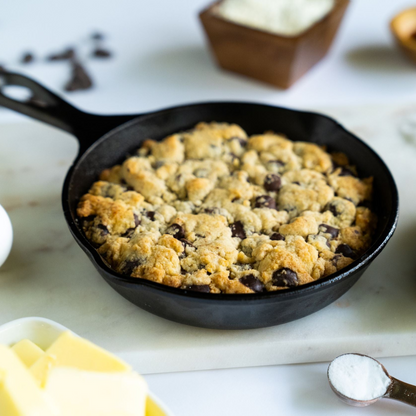 A freshly baked Sugar-Free Chocolate Chip Cookie from Lakanto sits in a black skillet on a marble surface. Nearby are ingredients like butter, almond flour, and sugar substitute, with chocolate chips scattered in the background.