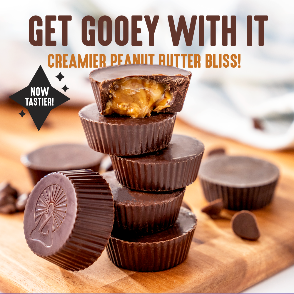 A stack of Dark Chocolate Peanut Butter Cups sits on a wooden board, one split open to show its gooey filling. Text reads Get Gooey With It: Creamier Peanut Butter Bliss! Now Tastier and Keto Friendly! Chocolate chips are scattered around.