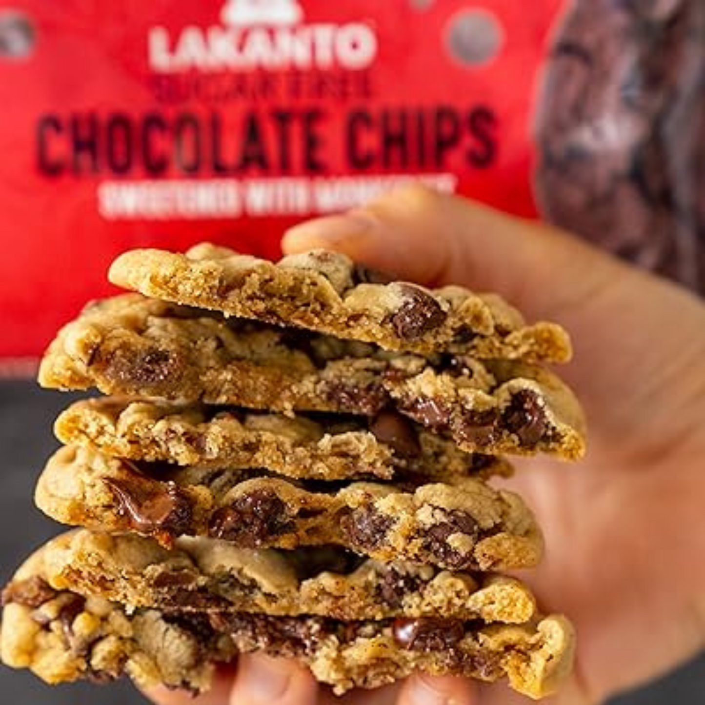 A hand holds a stack of chocolate chip cookies with gooey chocolate pieces. The blurred background package reads Lakanto Sugar-Free Chocolate Chips, sweetened with monk fruit, ideal for a keto-friendly treat.